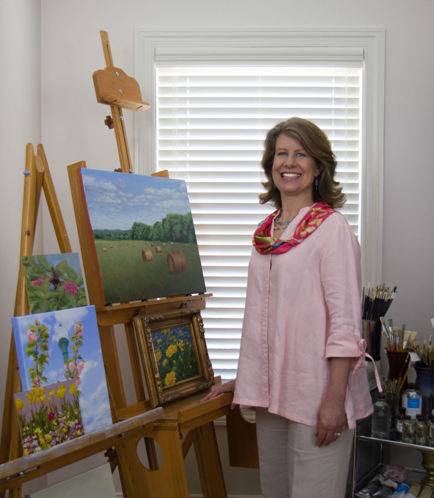 Renée Daly in her studio standing in front of two wooden easels displaying oil paintings. Brushes, paints and oils are on a shelf behind her.